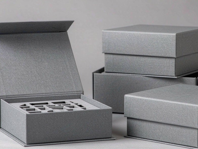 Blank greyboard two-piece rigid boxes: one open with the lid lifted off beside the base tray and die-cut insert, others closed showing the lid overhang seam